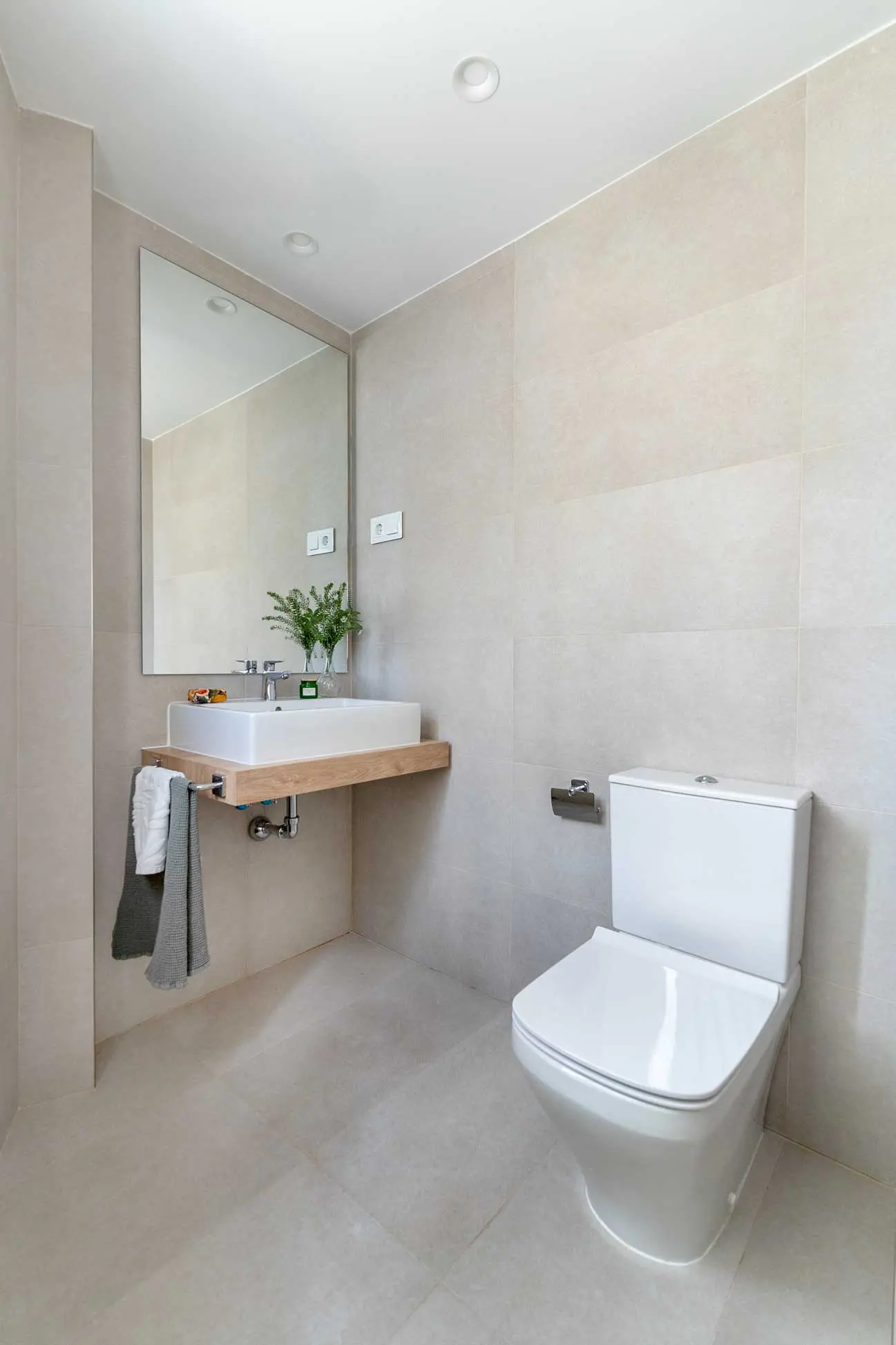 Aseo de cortesía con lavabo cuadrado sobre balda de madera, grifería cromada, espejo de gran tamaño e inodoro sobre paredes en tono crema.