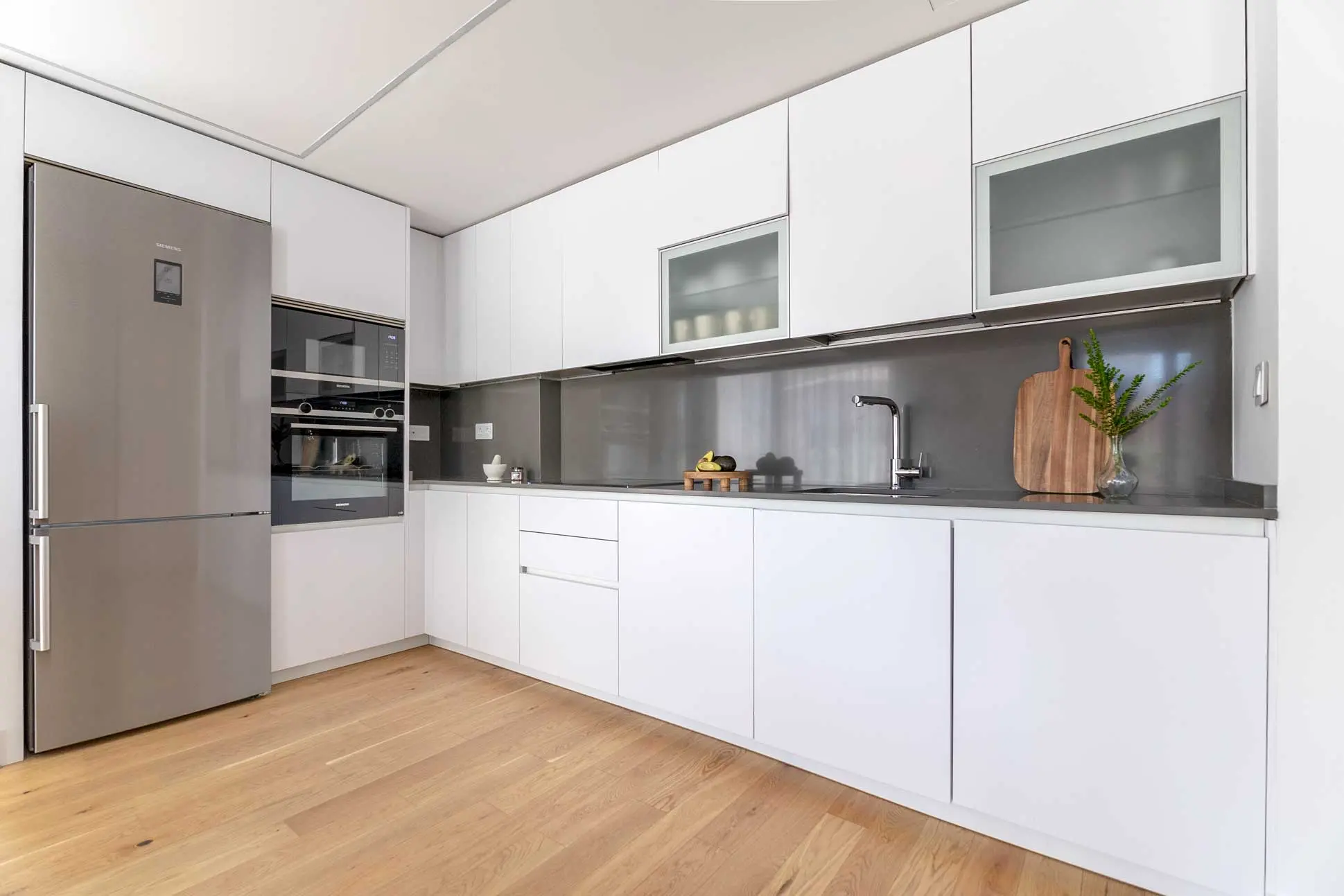 Cocina con frentes lacados en blanco mate, encimera y salpicadero en gris grafito, electrodomésticos integrados en acero inoxidable y suelo de madera clara.