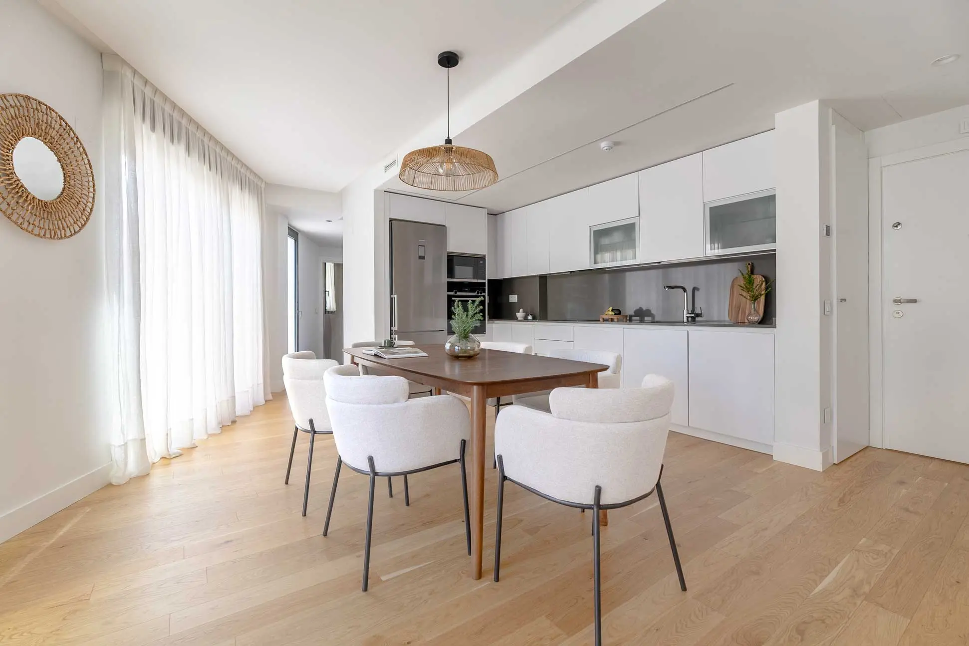 Comedor integrado con la cocina: mesa de madera, sillones tapizados en crema con patas negras, lámpara colgante de ratán trenzado y espejo redondo trenzado.