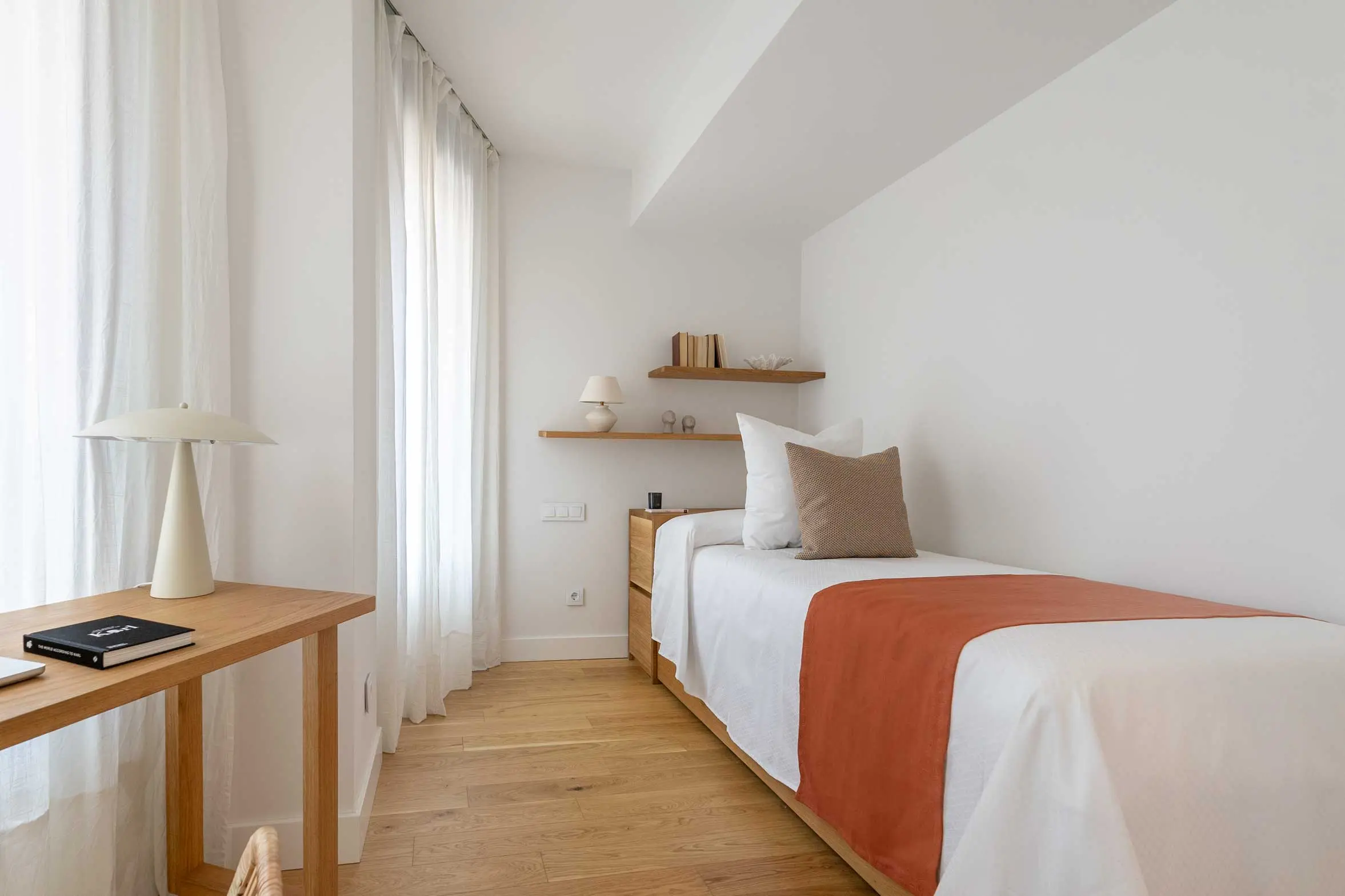 Dormitorio individual con cama vestida con un pie de cama color teja, estantes volados de madera, escritorio junto a la ventana y lámpara de sobremesa cónica.
