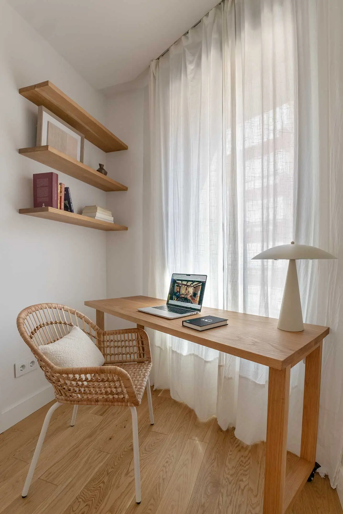 Rincón de estudio junto al ventanal, con escritorio de madera, silla de mimbre, estantes volados y una lámpara de mesa de diseño.