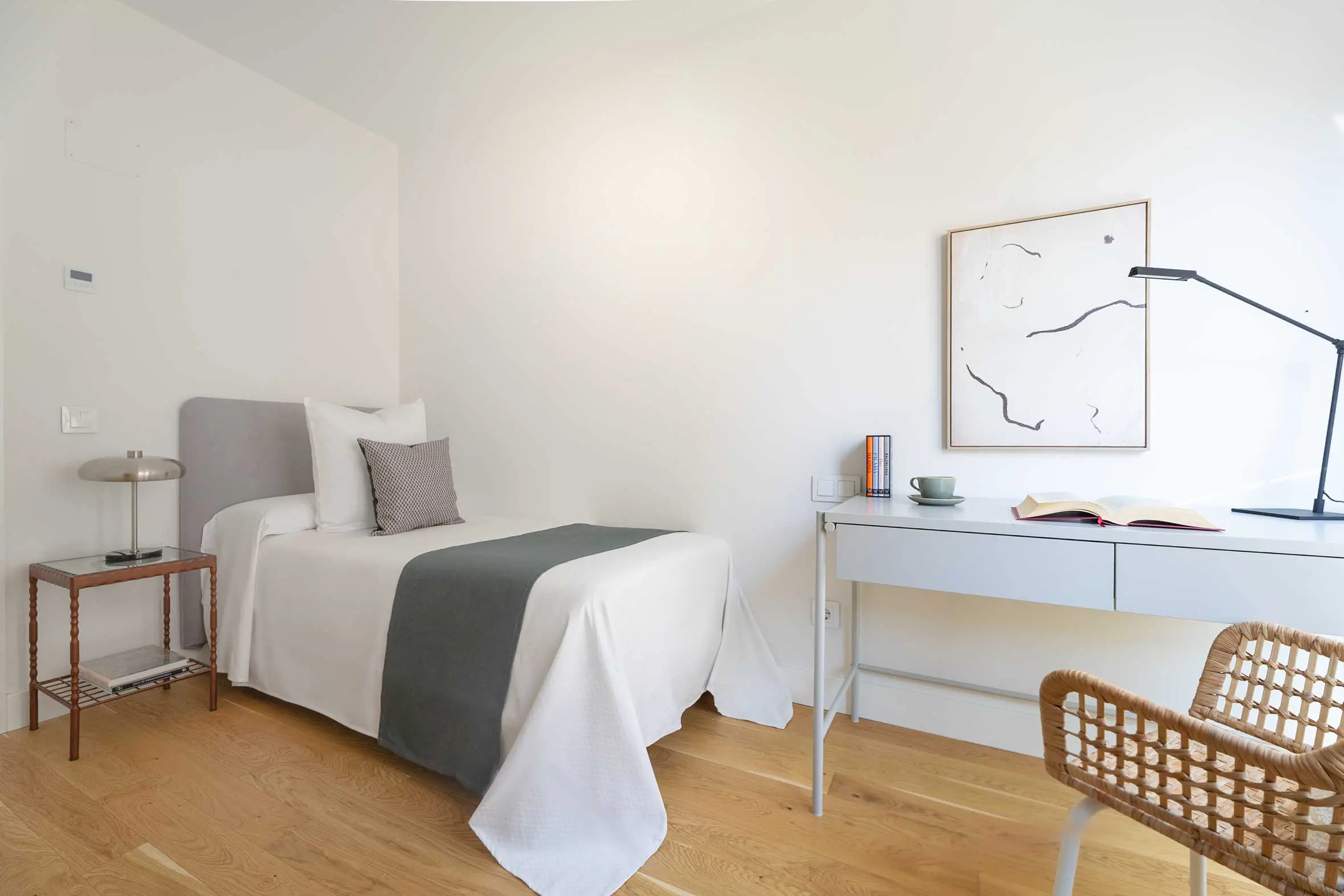 Segundo dormitorio individual con cama de pie de cama gris, mesilla clásica de madera, escritorio blanco y una lámina abstracta en la pared.