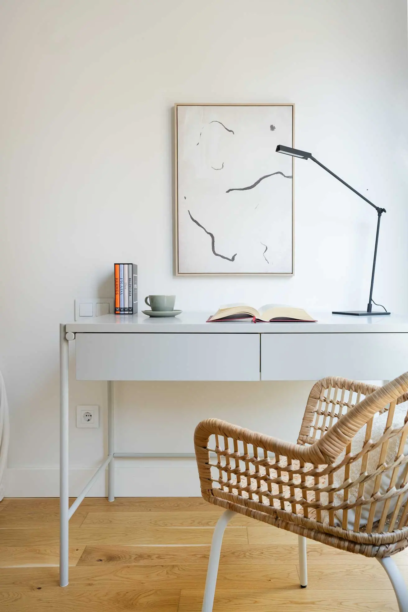 Detalle de la zona de estudio con escritorio blanco, flexo de diseño negro, un libro abierto y silla de mimbre.
