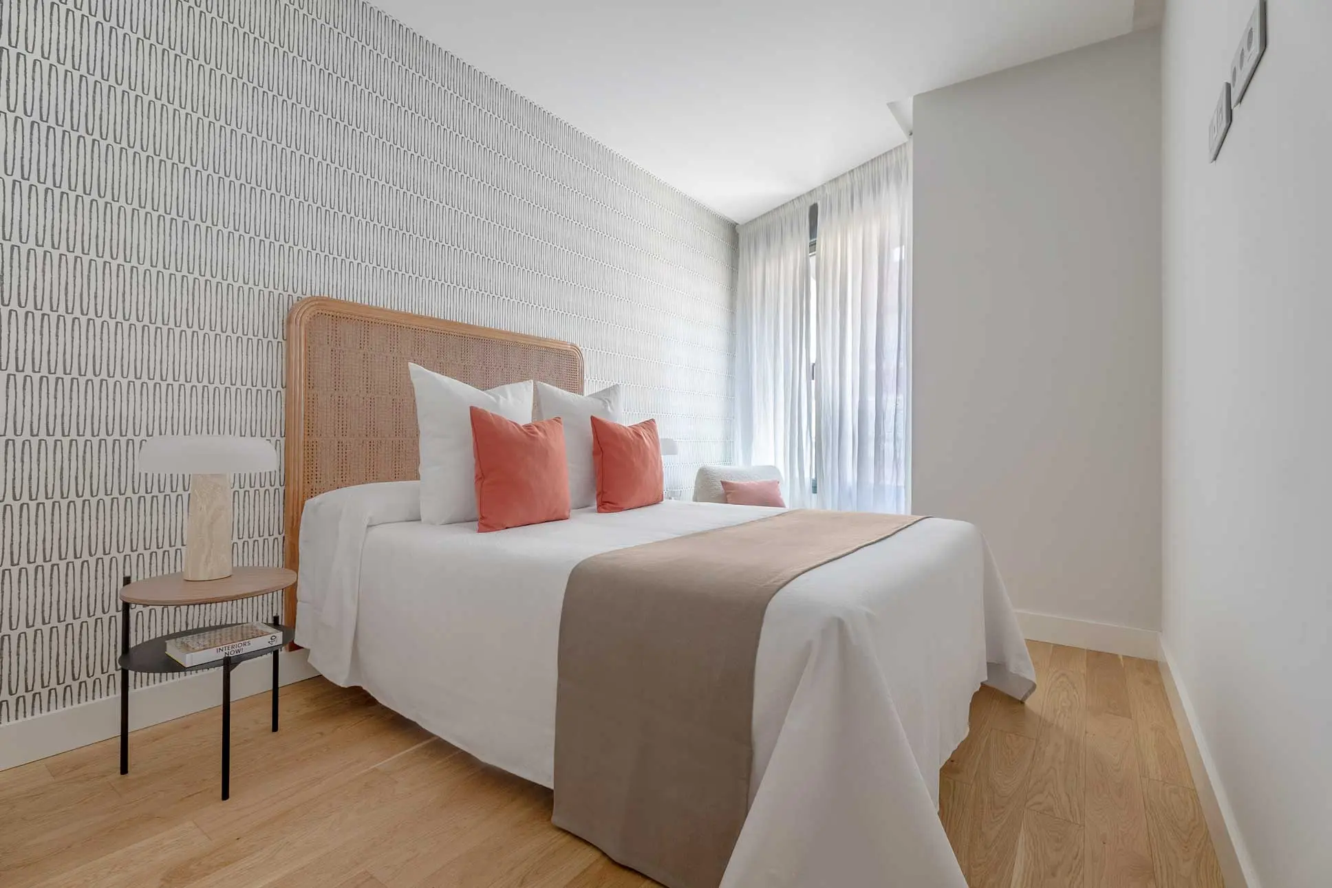 Dormitorio principal con cama doble, cojines coral, cabecero de rejilla de ratán y una pared empapelada con un sutil patrón geométrico lineal.