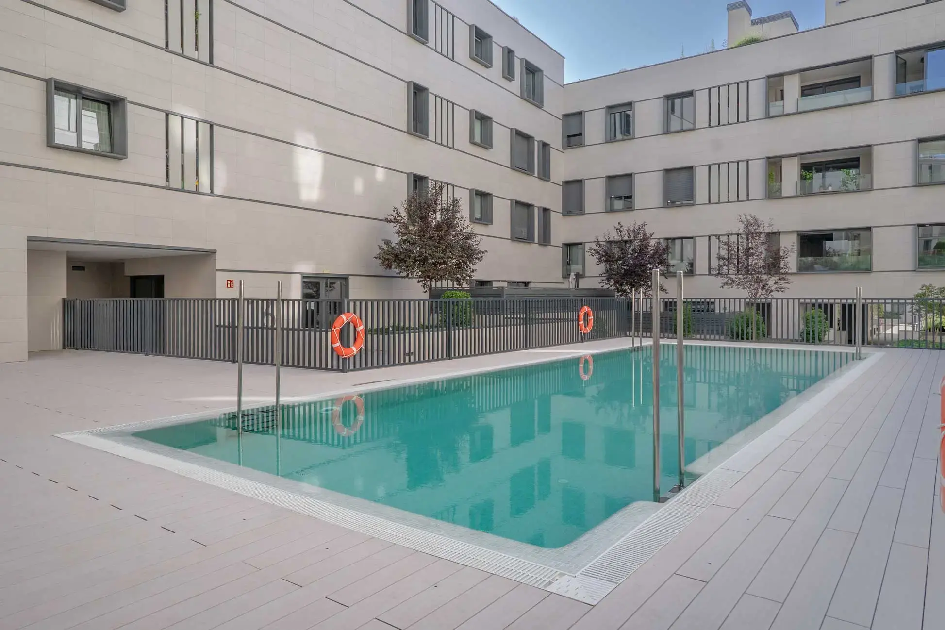 Piscina alargada y vallada en el patio interior del complejo residencial, con fachadas grises y salvavidas naranjas.