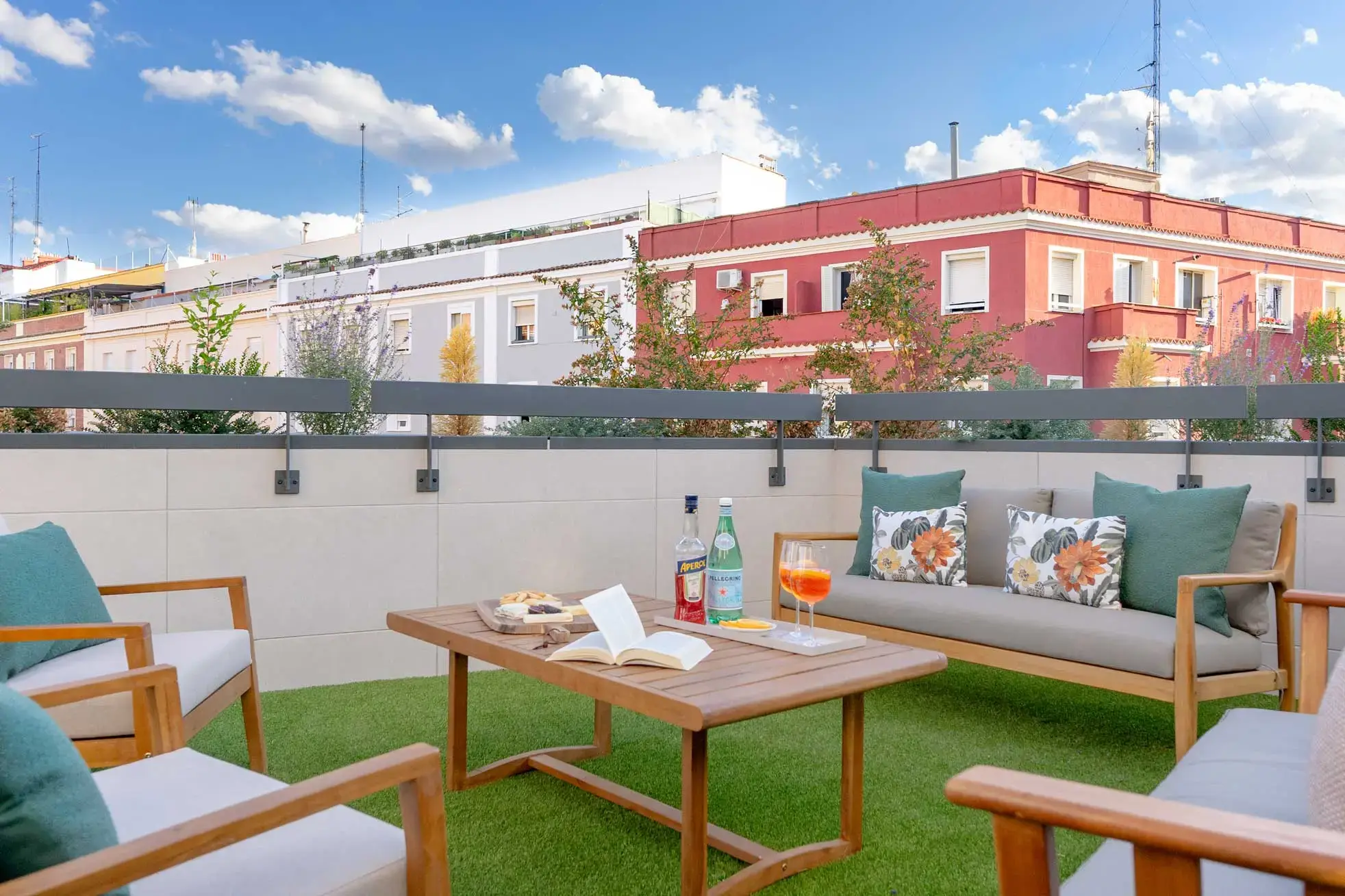 Área de descanso en una terraza de ático con césped artificial, sofás con estructura de madera, cojines estampados con motivos florales y una mesa de centro baja con bebidas frente a edificios urbanos.
