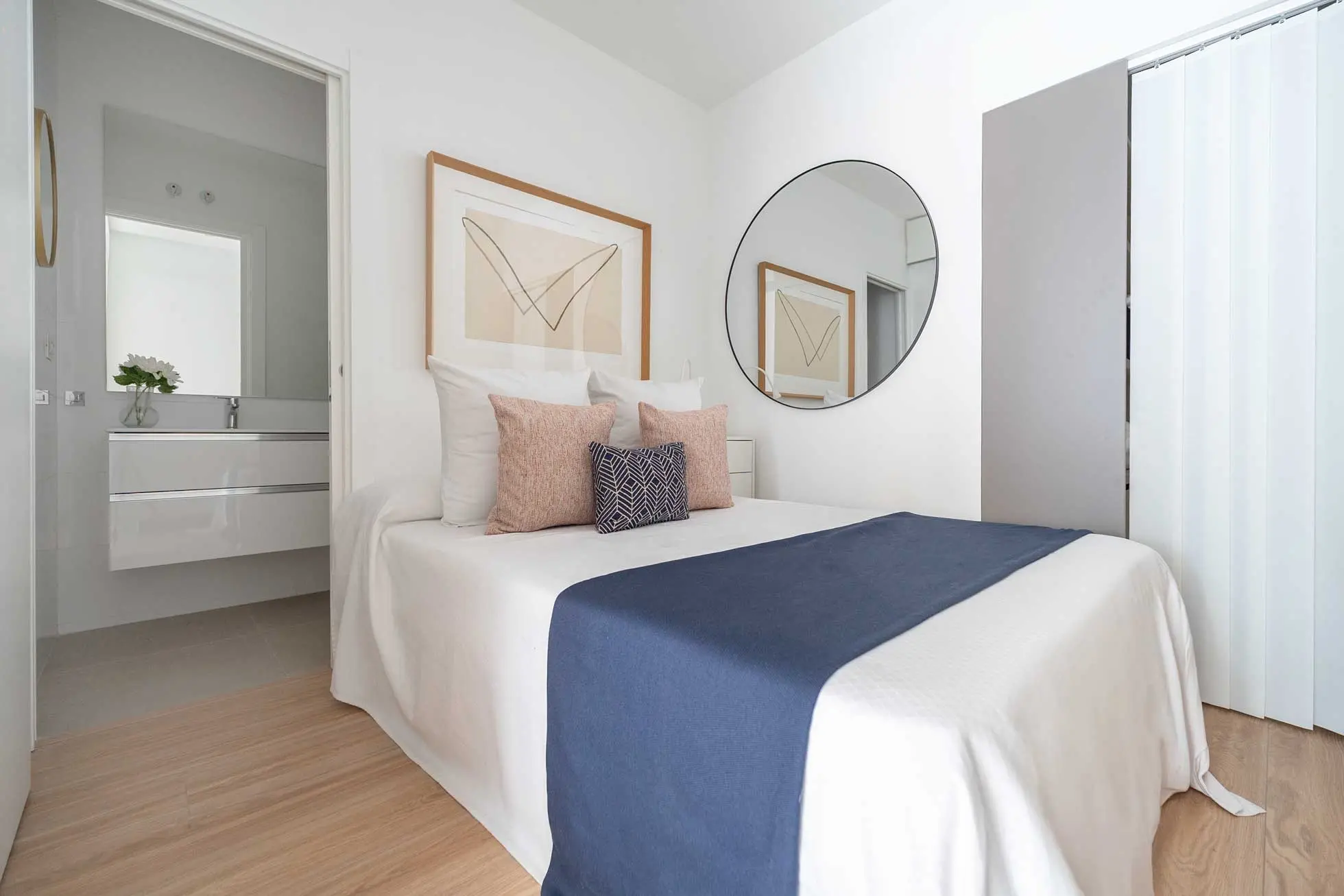 Dormitorio principal con cama doble vestida en tonos blancos y azul marino, mesilla minimalista y acceso directo al baño integrado