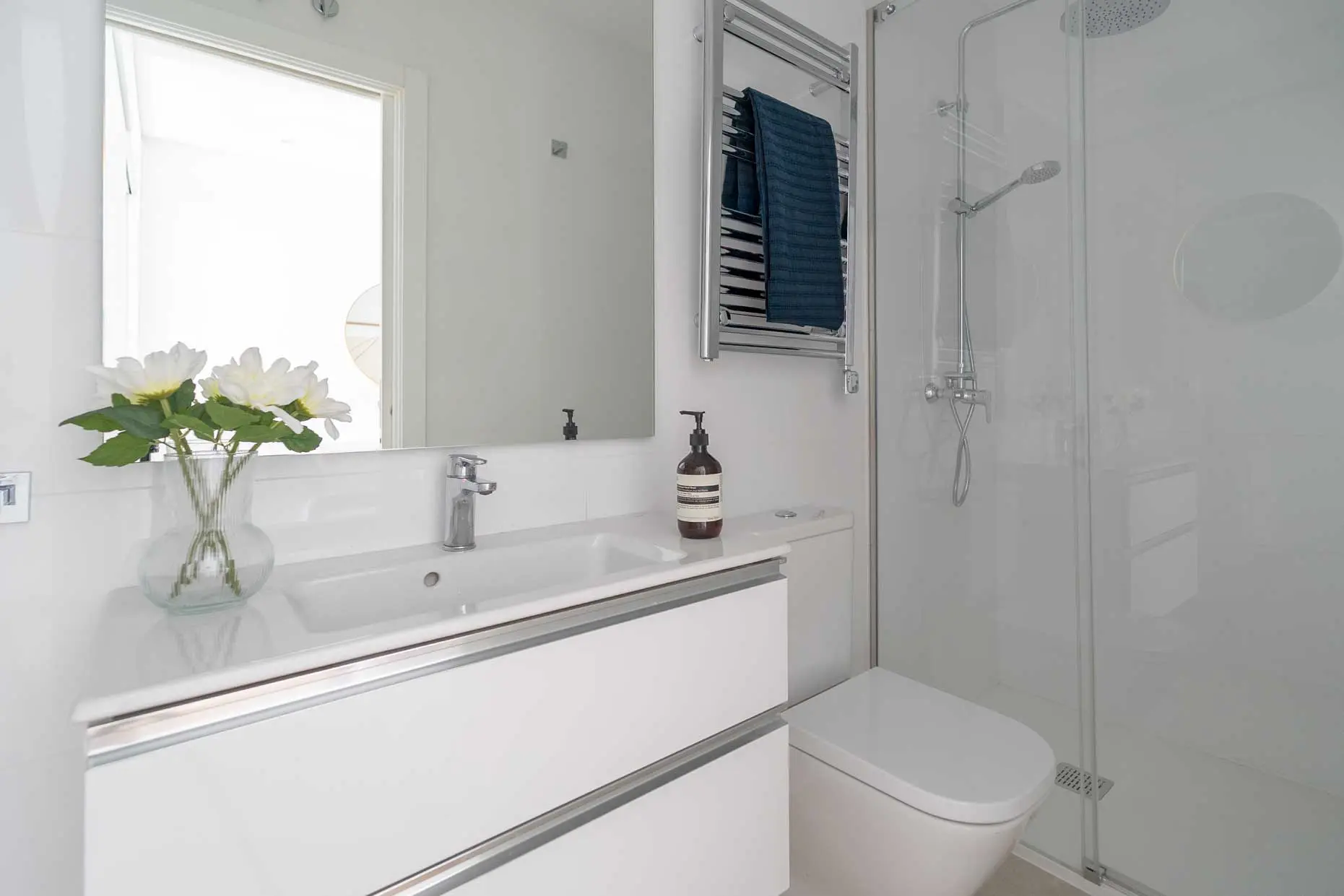 Detalle del lavabo moderno con grifería cromada, jarrón con flores blancas decorativas y un radiador toallero de diseño