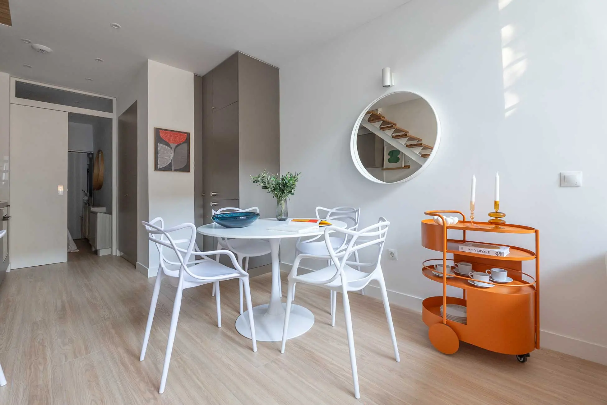 Zona de comedor con mesa redonda blanca, sillas de diseño entrelazadas y el carrito auxiliar caldera bajo el gran espejo circular