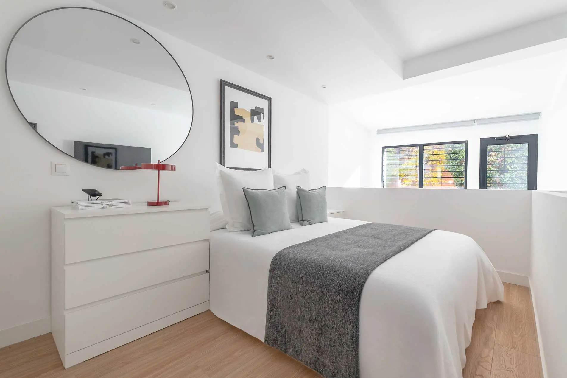 Dormitorio en la planta alta tipo loft con cama doble, camino gris, cojines verde salvia y una cómoda blanca frente a un espejo maxi