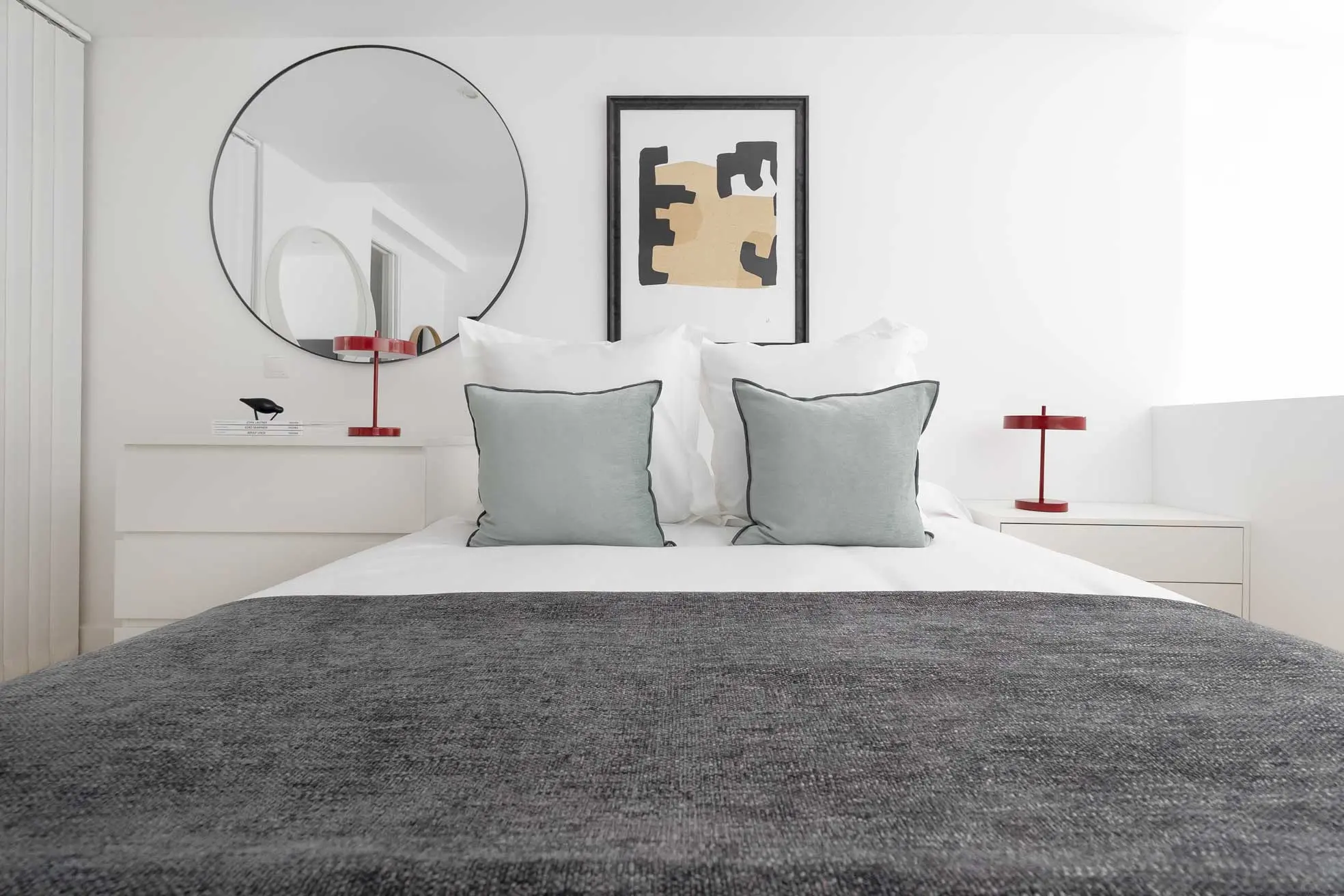 Plano medio frontal de la cama del dormitorio superior con almohadones texturizados grisáceos, lámparas de mesa rojas y cuadro de arte moderno
