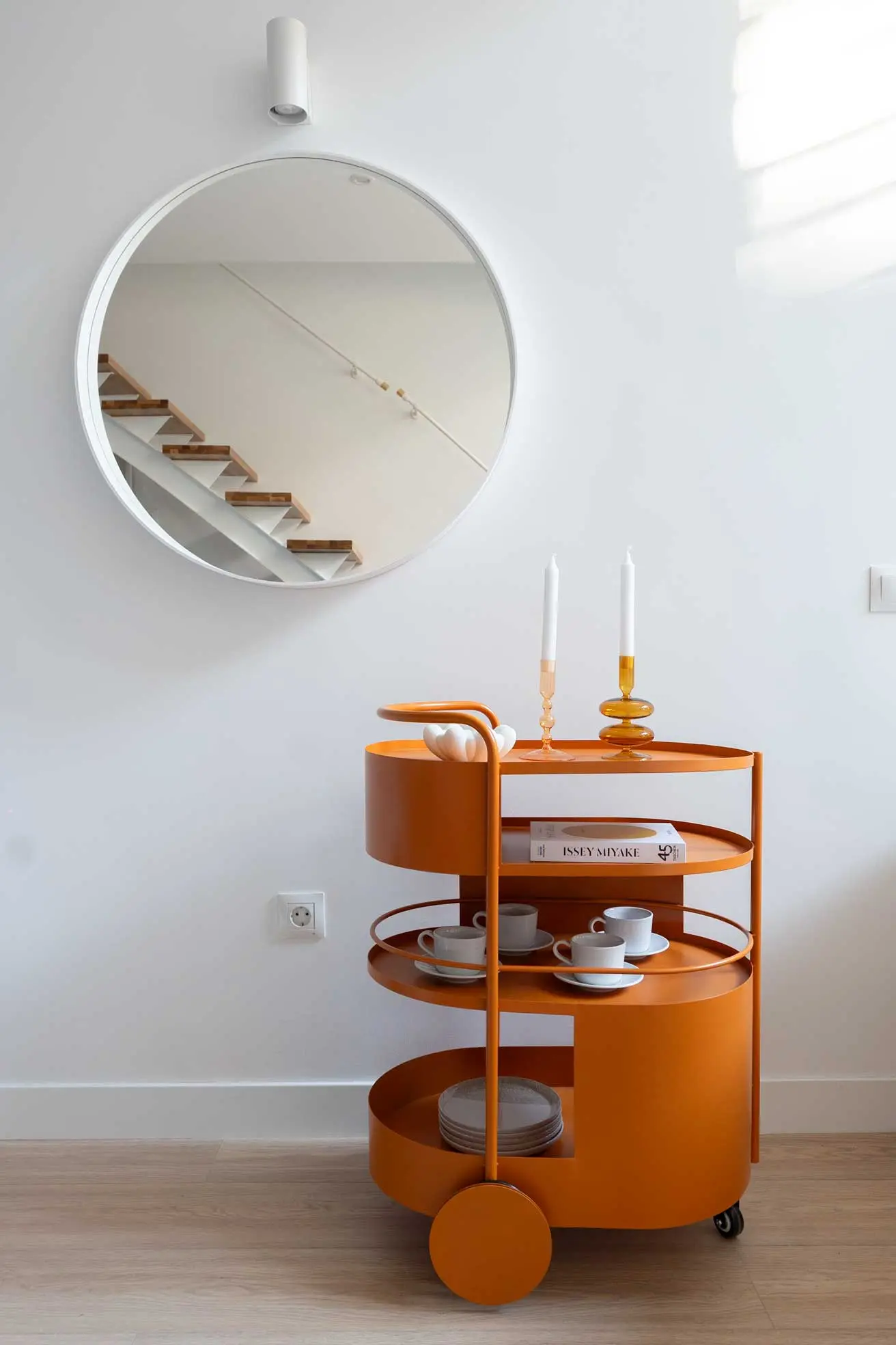 Detalle de estilismo con un carrito camarera de diseño color naranja caldero con tazas y velas frente a un espejo redondo que refleja una escalera calada
