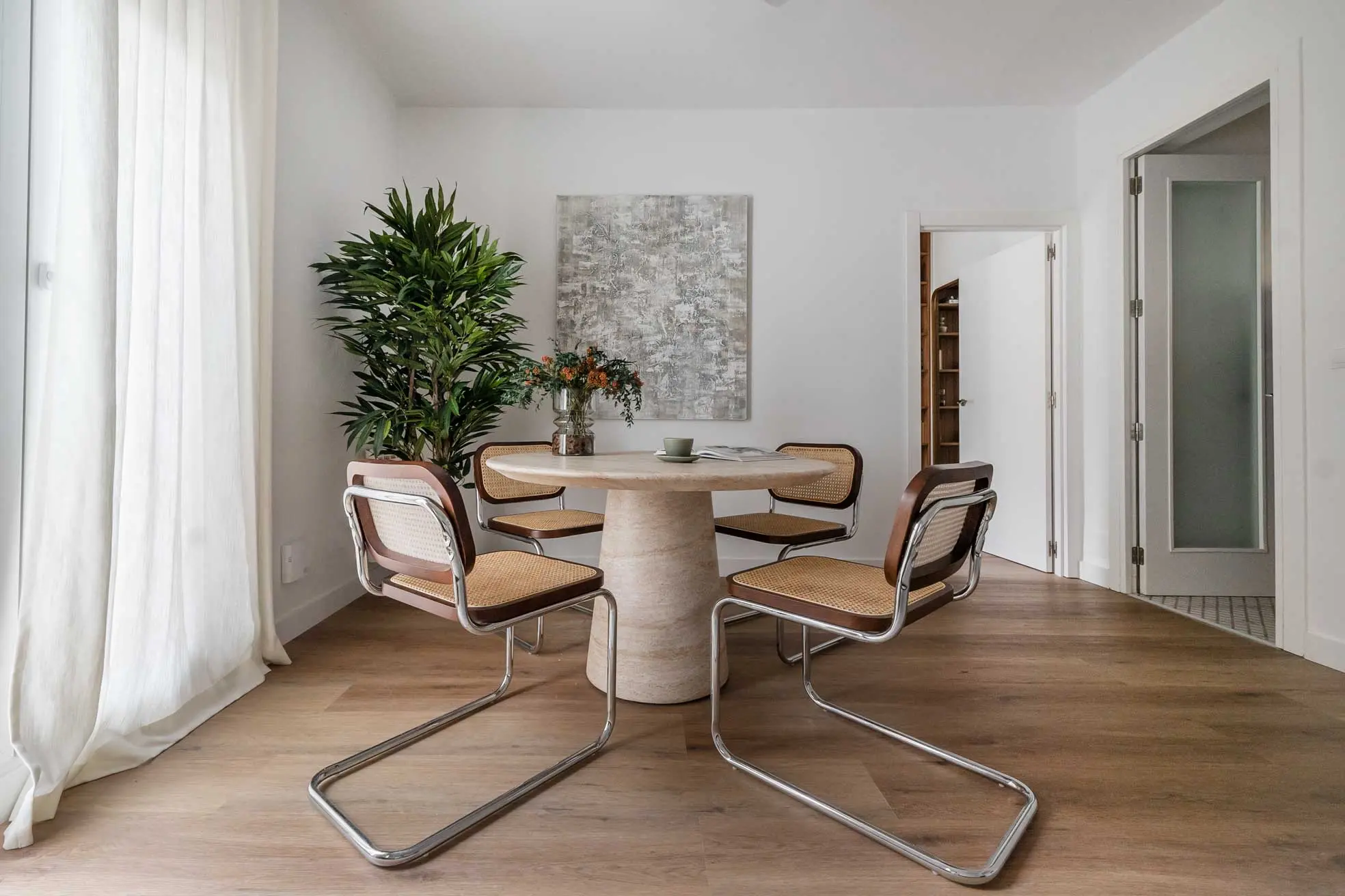 Zona de comedor de diseño con mesa redonda de piedra travertino, sillas estilo Cesca de rejilla y un gran cuadro abstracto