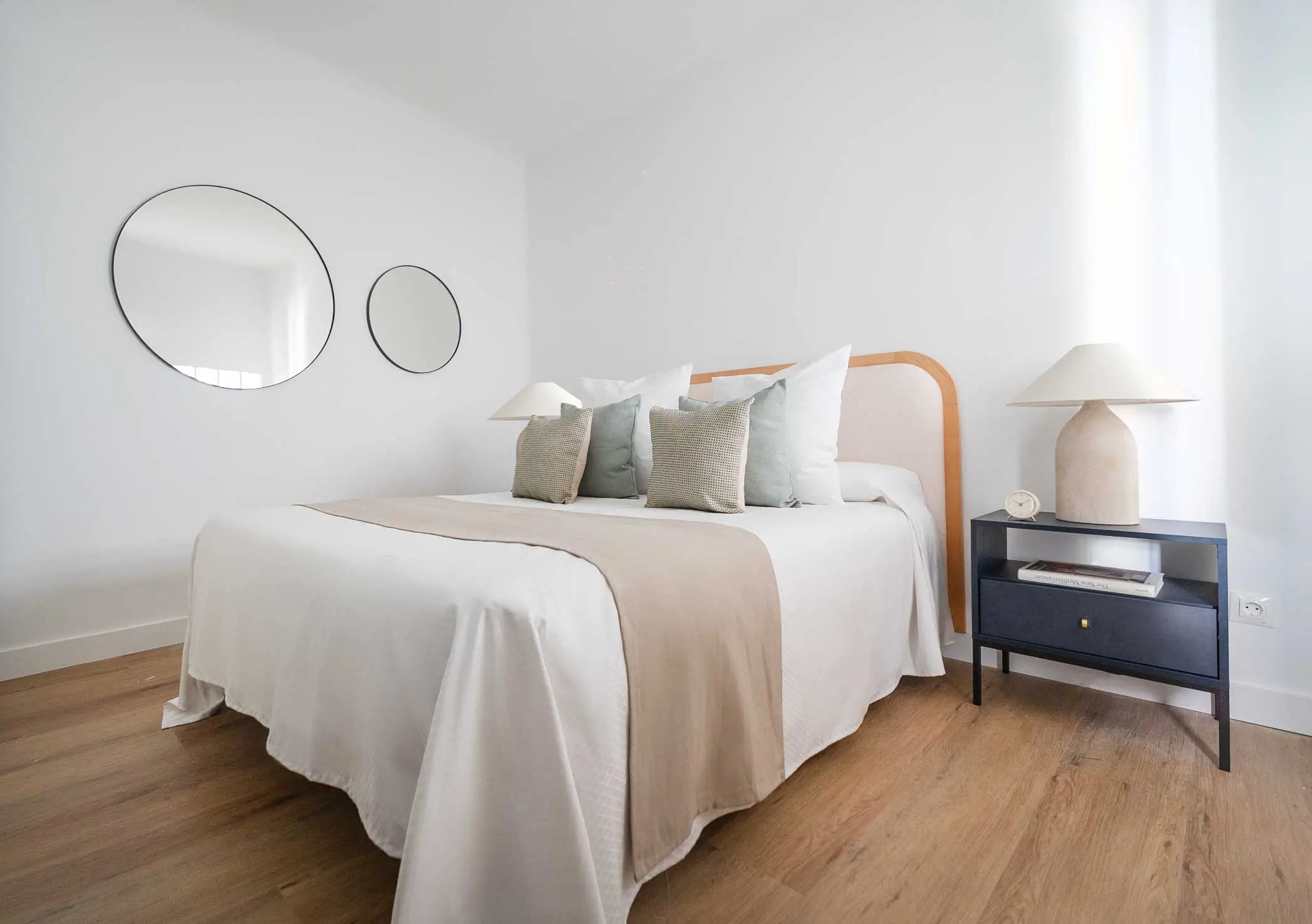 Dormitorio principal con cama doble perfectamente vestida, cojines en tonos verde salvia, espejos circulares y mesilla de noche minimalista