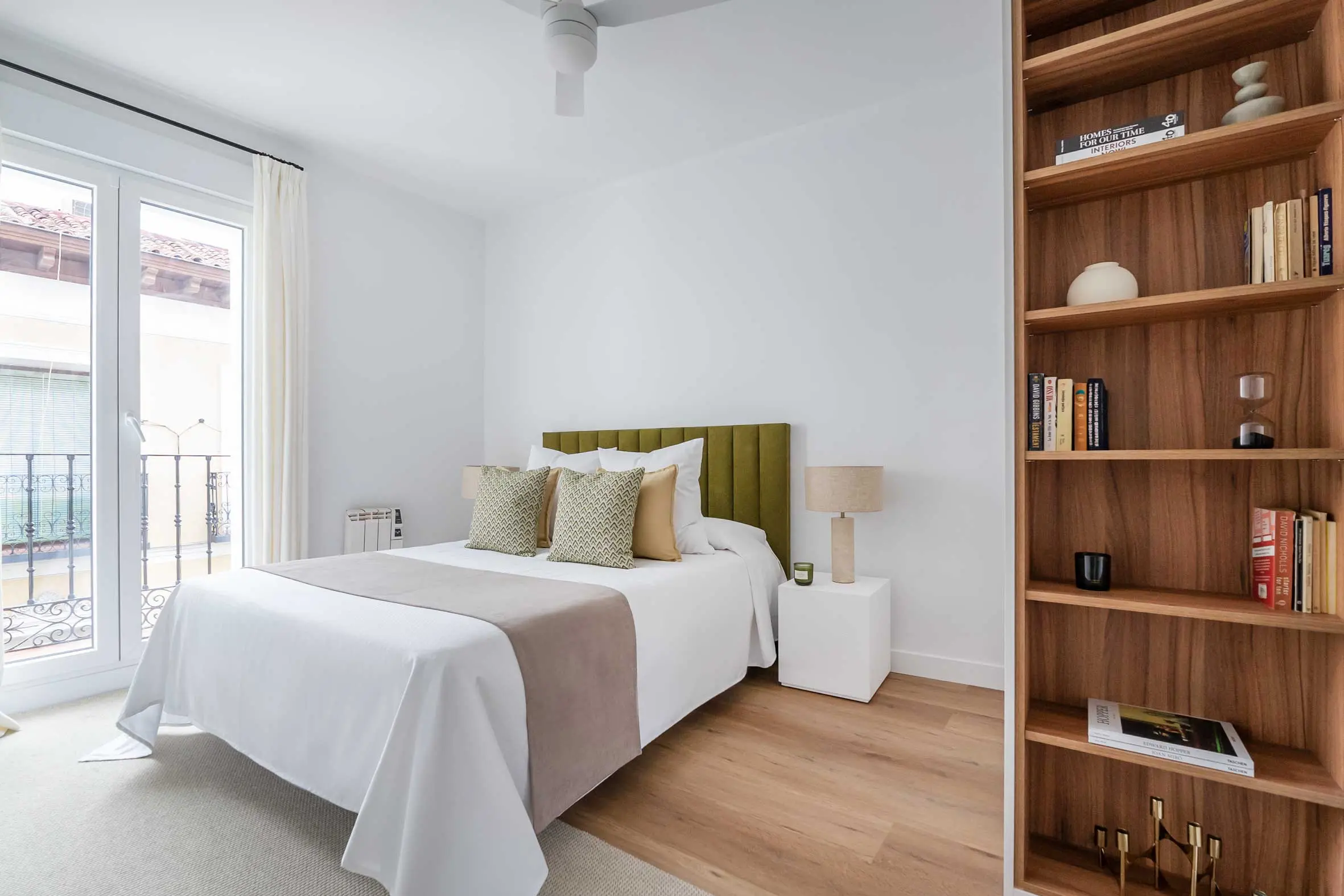 Perspectiva luminosa del dormitorio principal con un gran mueble de estantería de madera en primer plano, cama doble con cabecero verde y balcón tradicional al fondo