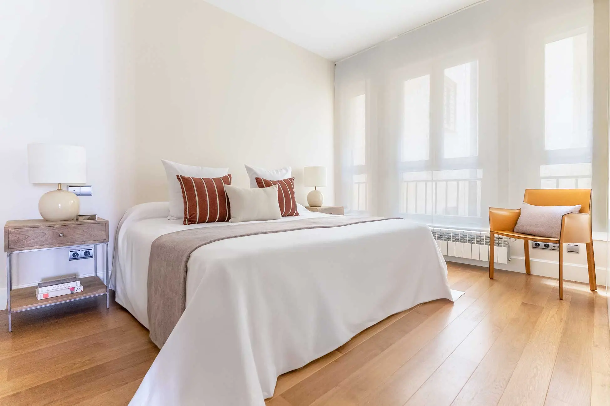 Dormitorio principal con una amplia cama de matrimonio vestida minuciosamente con cojines de rayas cobrizas y butaca de cuero