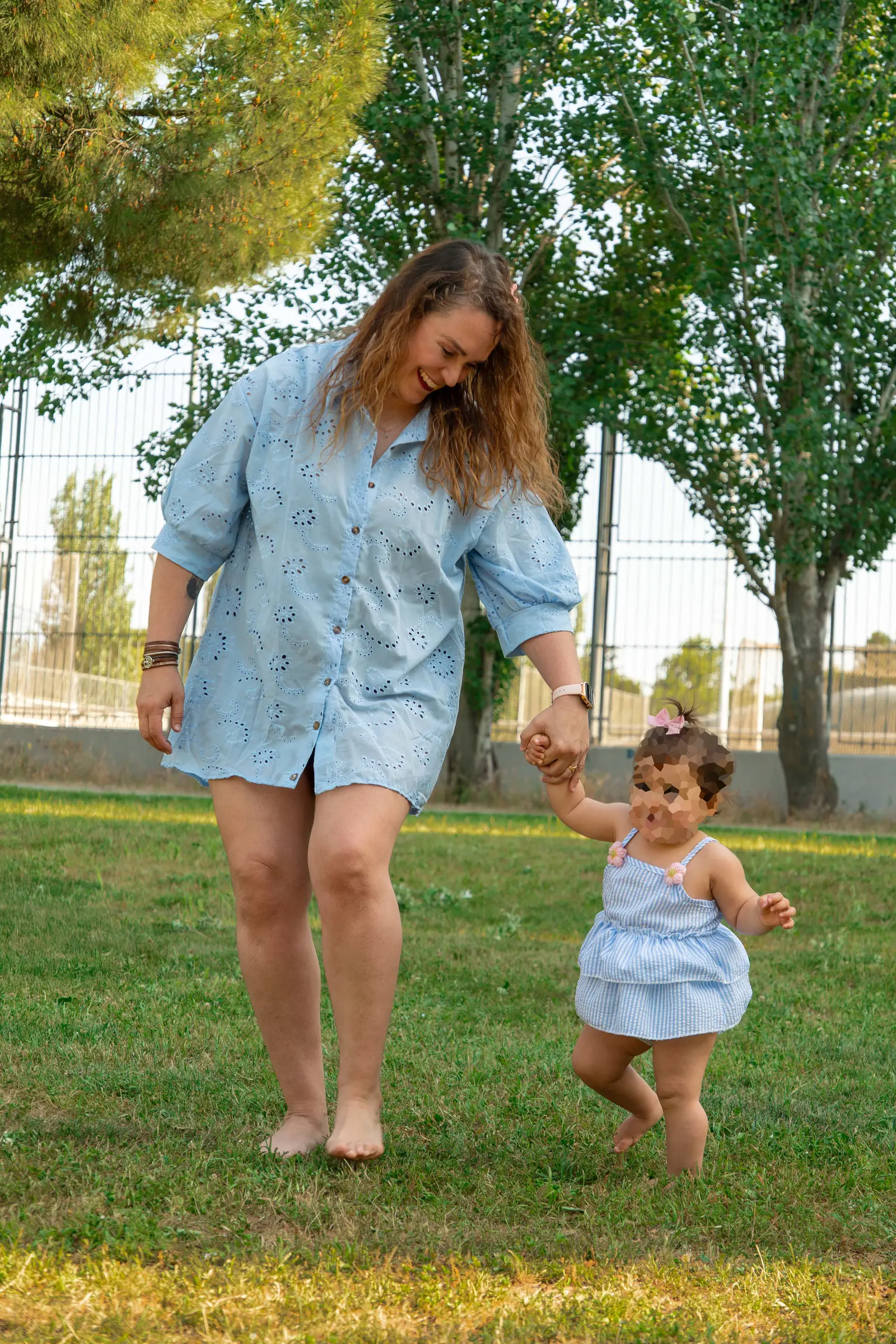 Padre con camisa rosa, descalzo sobre la hierba, caminando de la mano de su hija pequeña entre árboles.