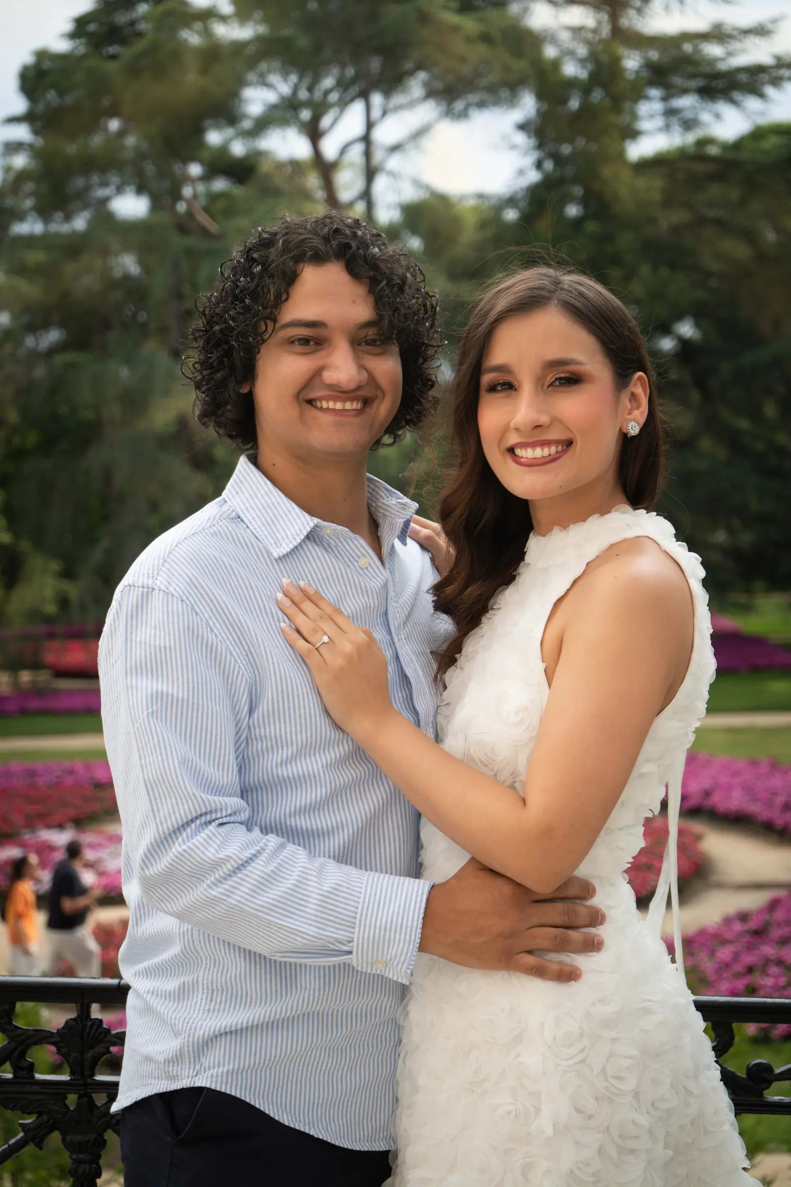 Retrato de cuerpo entero de la pareja abrazada en un sendero arbolado; ella, con vestido blanco corto, mira a cámara y él la mira a ella.
