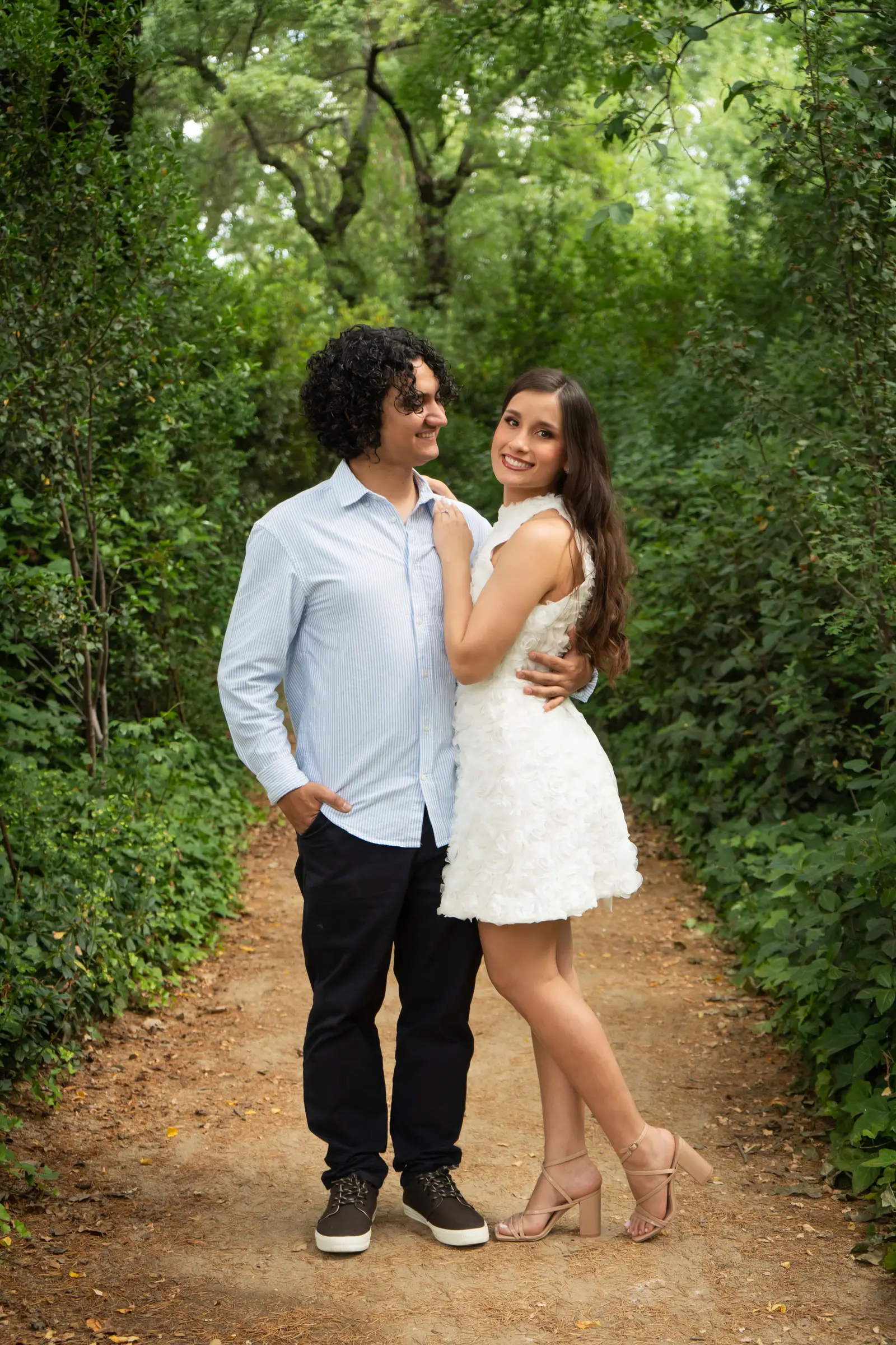La pareja de pie en un sendero del parque; ella apoya la mano sobre el pecho de él, con el anillo visible, y ambos sonríen a cámara.