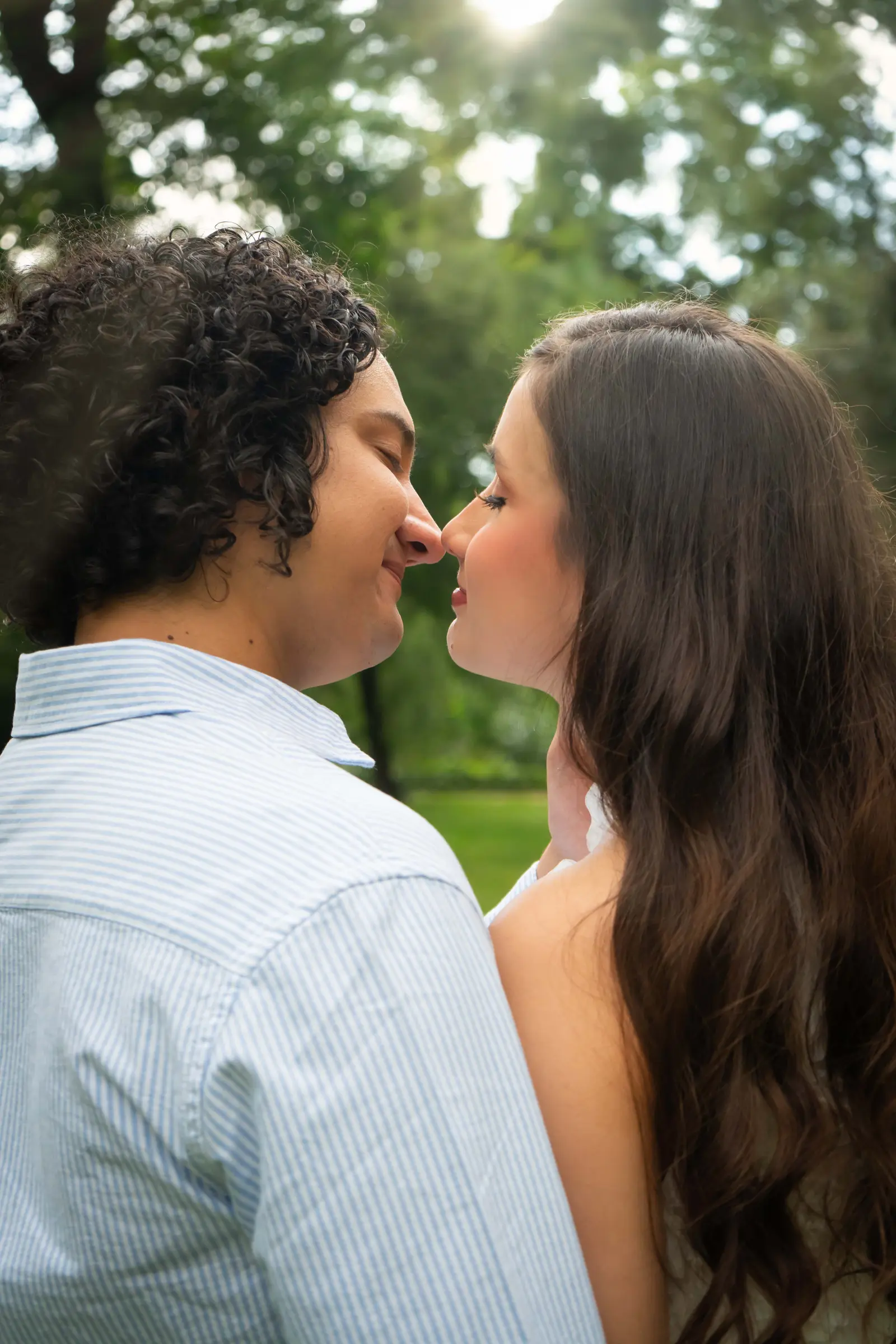Perfil de la pareja a punto de besarse a contraluz, con la espalda de él en primer plano y el sol entre los árboles.