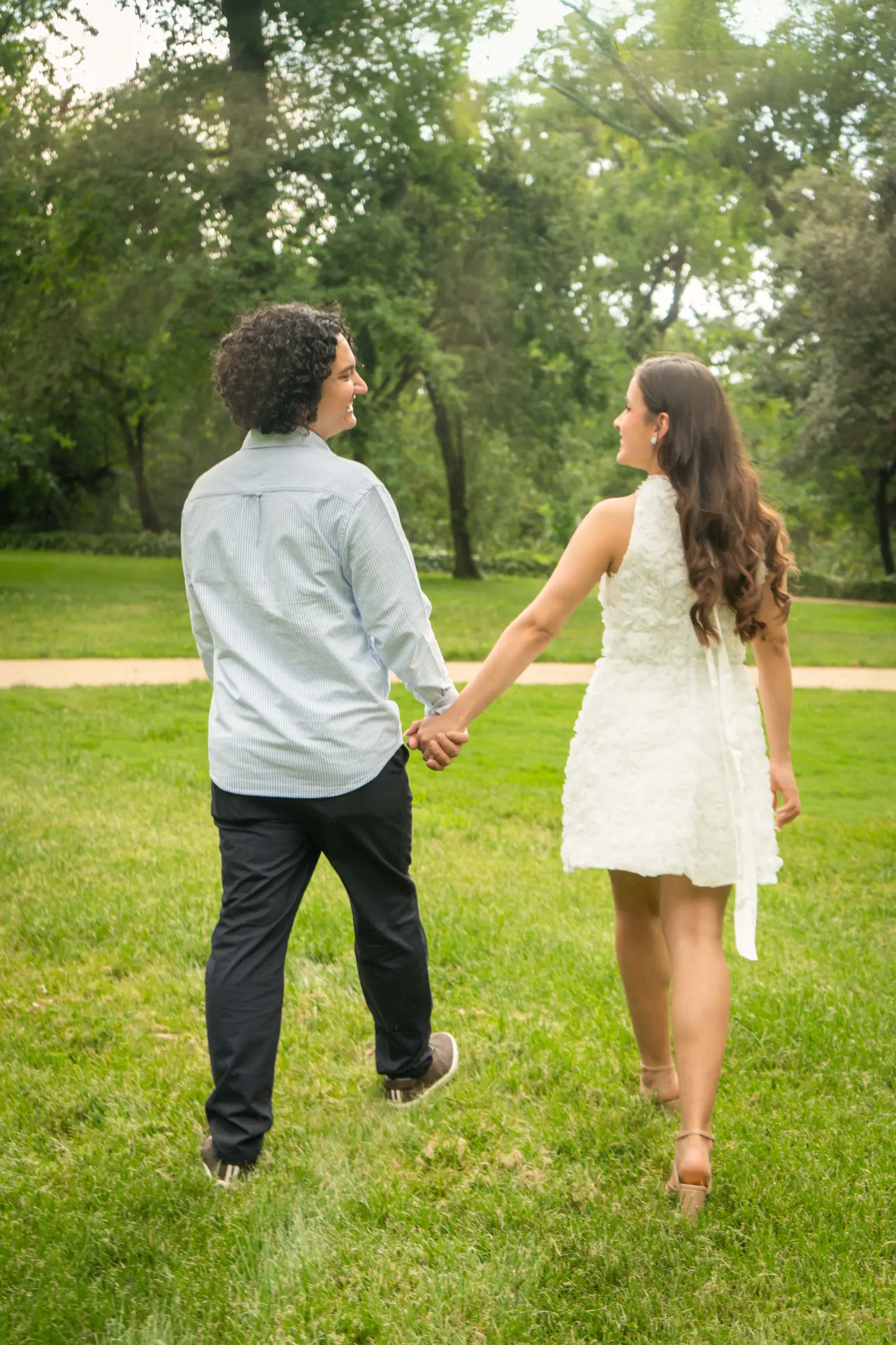 La pareja camina de la mano y de espaldas sobre el césped de un parque, mirándose con complicidad.