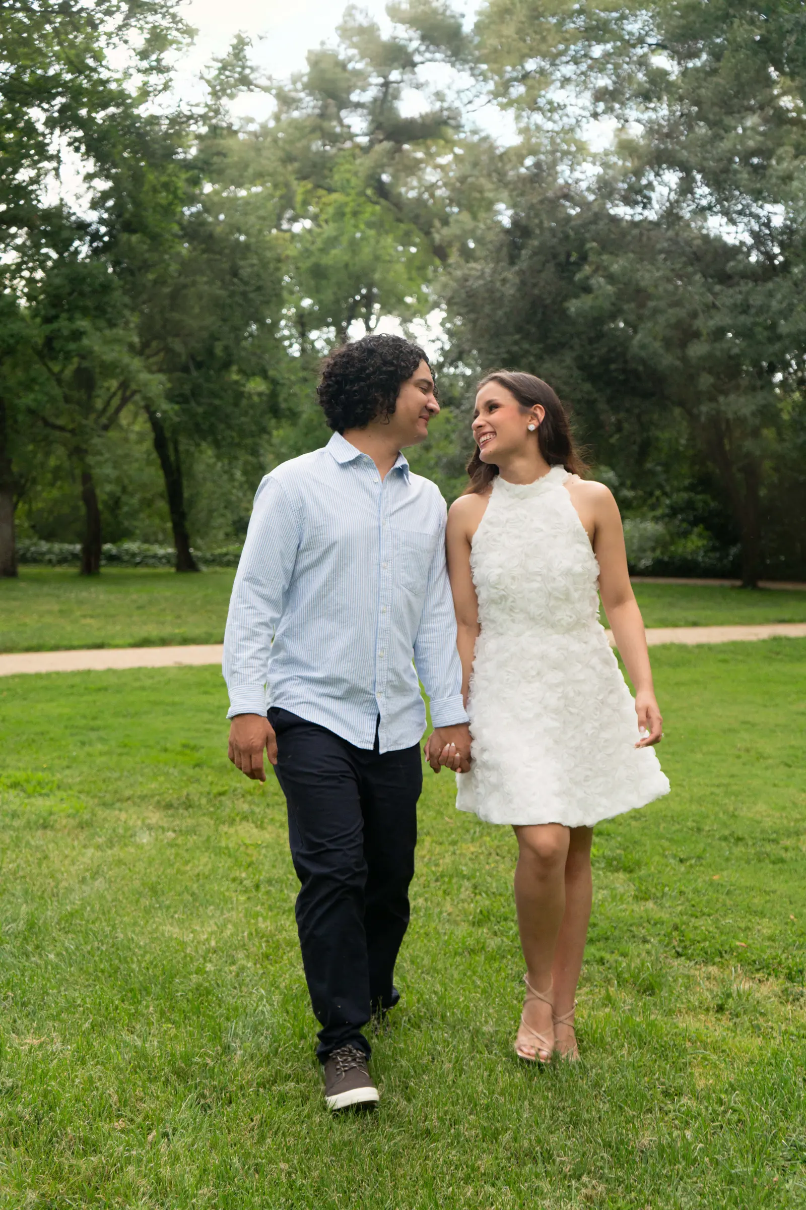 La pareja camina de la mano hacia la cámara sobre el césped, mirándose y sonriendo entre grandes árboles.