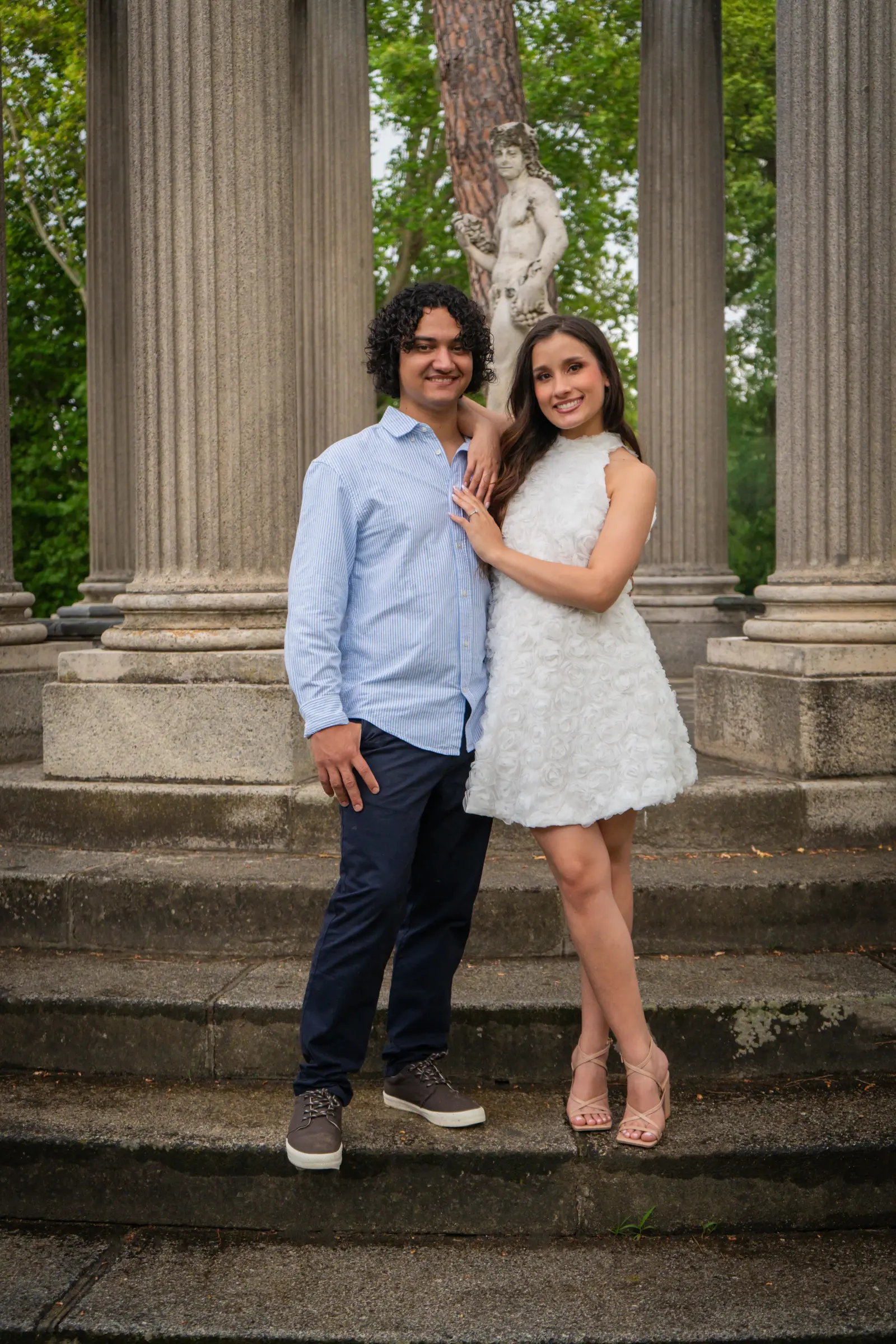 Retrato de la pareja de pie sobre unas escaleras de piedra frente a imponentes columnas clásicas, con una estatua al fondo.