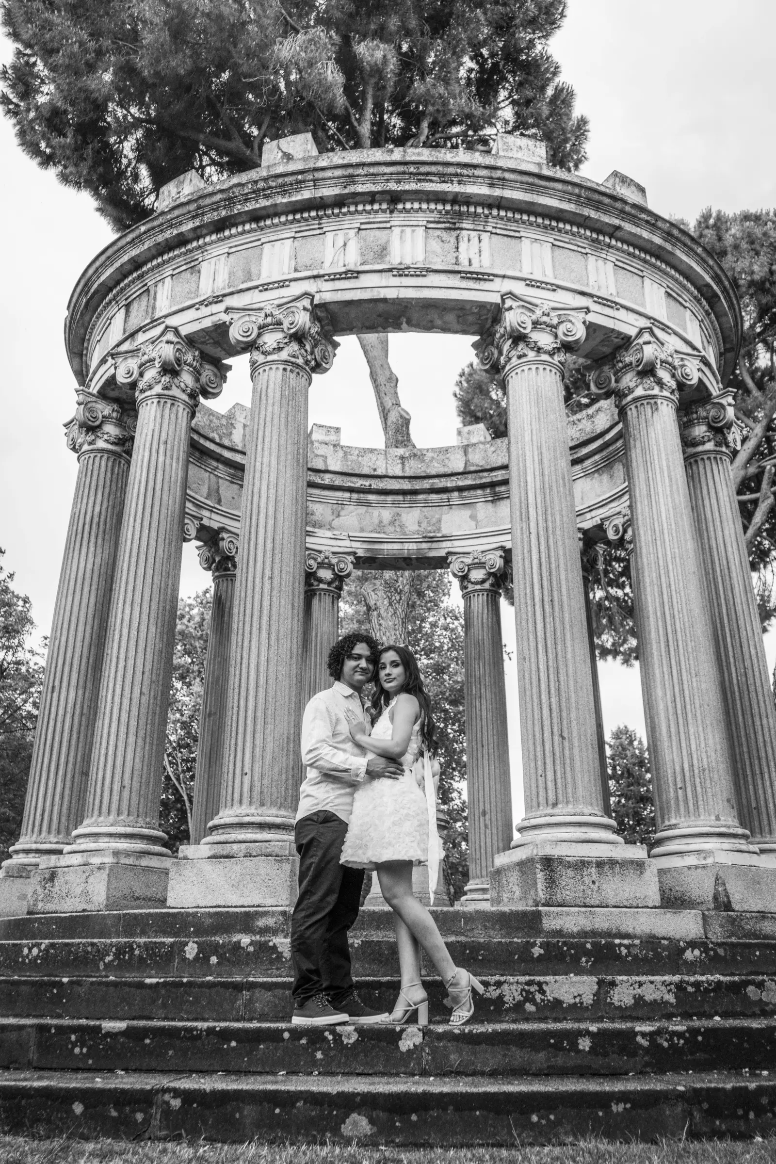 Fotografía artística en blanco y negro de la pareja abrazada bajo un templete circular de columnas de estilo clásico.