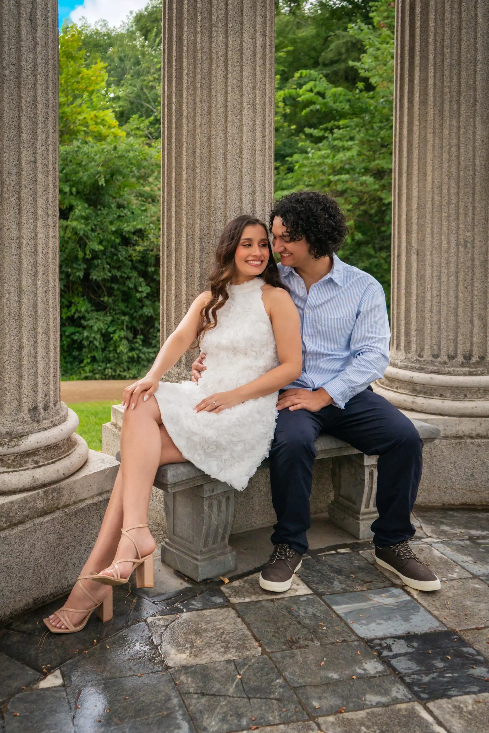 La pareja sentada en un banco de piedra entre grandes columnas, mirándose con complicidad sobre un suelo empedrado.