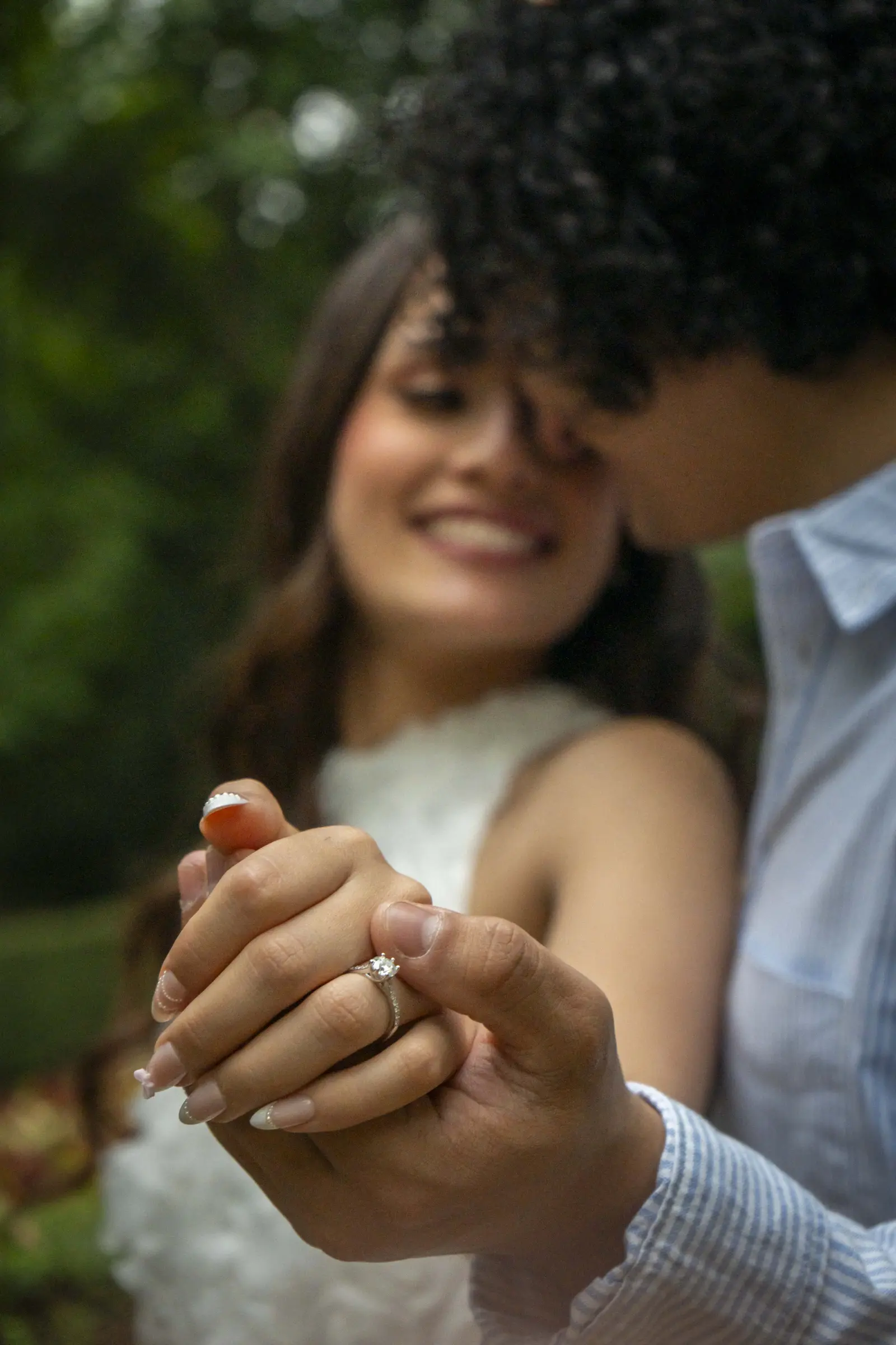 Primer plano de las manos entrelazadas de la pareja con un anillo de compromiso enfocado en el dedo de ella; al fondo, desenfocados, ambos sonríen muy juntos.